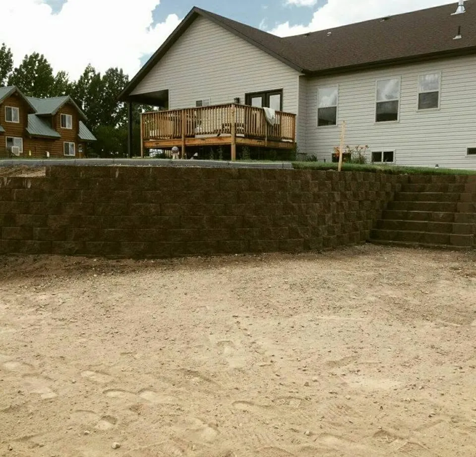 Long brown block retaining wall with built-in block steps below a white-sided house with a wood deck.