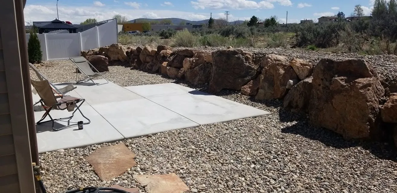 Row of large reddish boulders forming a low wall behind a new concrete patio pad with river rock and flagstone steppers.