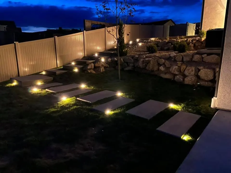 Concrete stepping pads crossing a lawn up to a boulder retaining wall, lit by small ground lights at night.