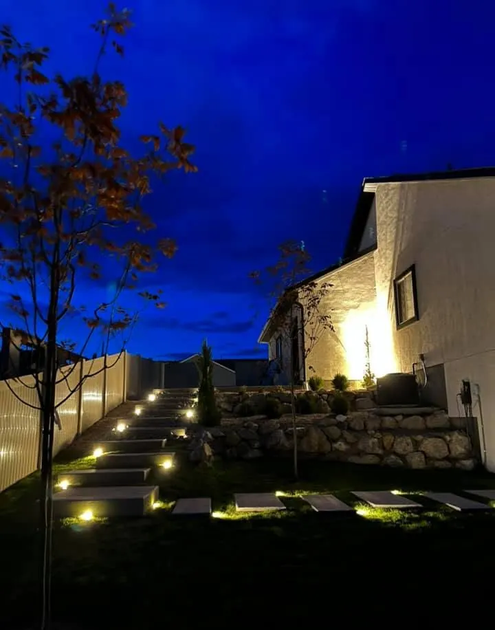Concrete step pads in a lawn and a stepped path up a slope, each lit by in-ground lights, beside a boulder wall.