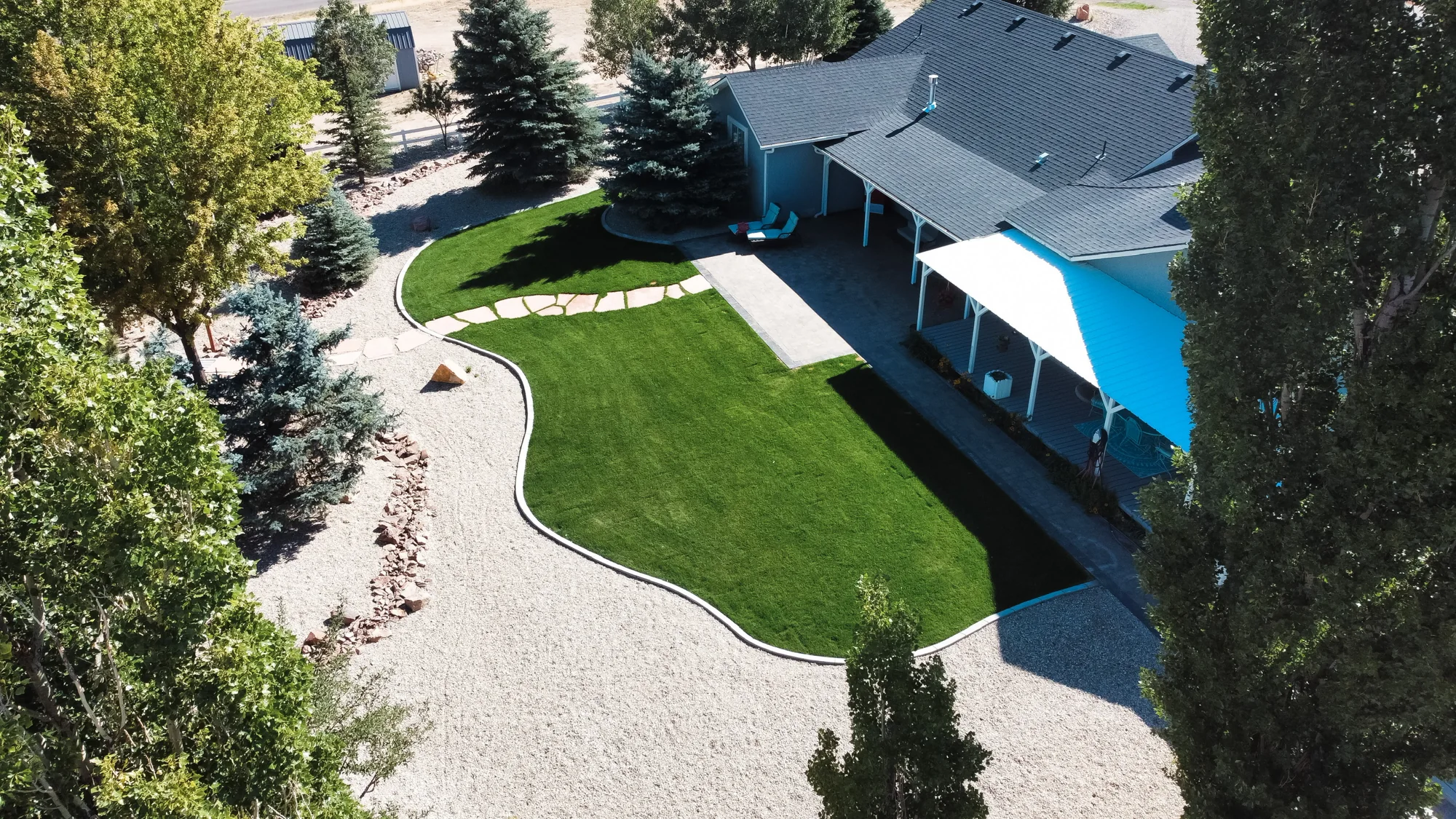 Aerial view of a green backyard lawn with curved gravel borders, trees and a covered patio.