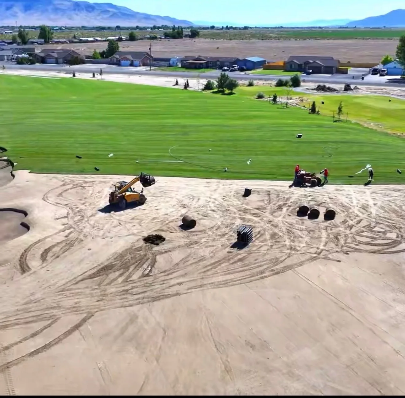 Wide view of green turf adjoining a dirt work area with machines and crew members.