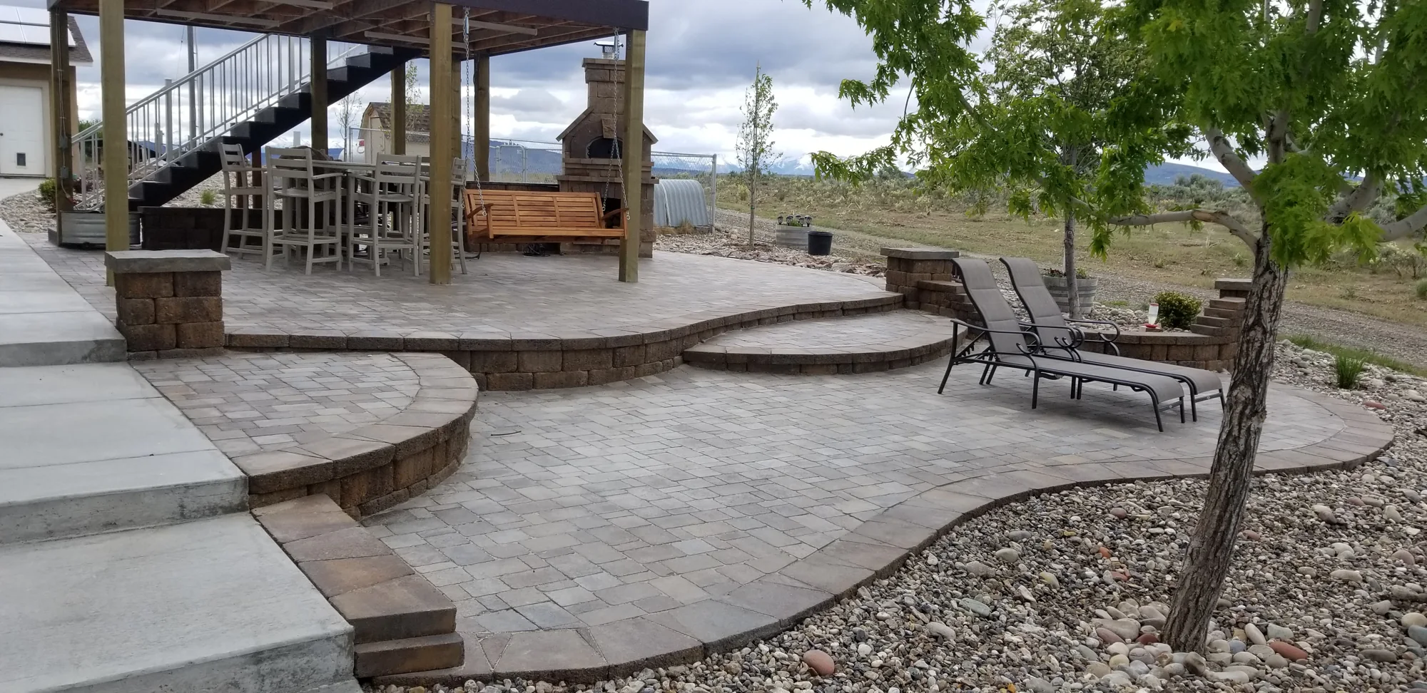 Paver patio with a covered outdoor kitchen, seating, a mature tree and rock landscaping.